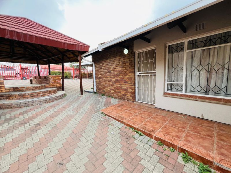 Rear exterior view featuring a paved courtyard, a covered gazebo with brick seating, and a tiled patio area adjacent to the main house structure.
