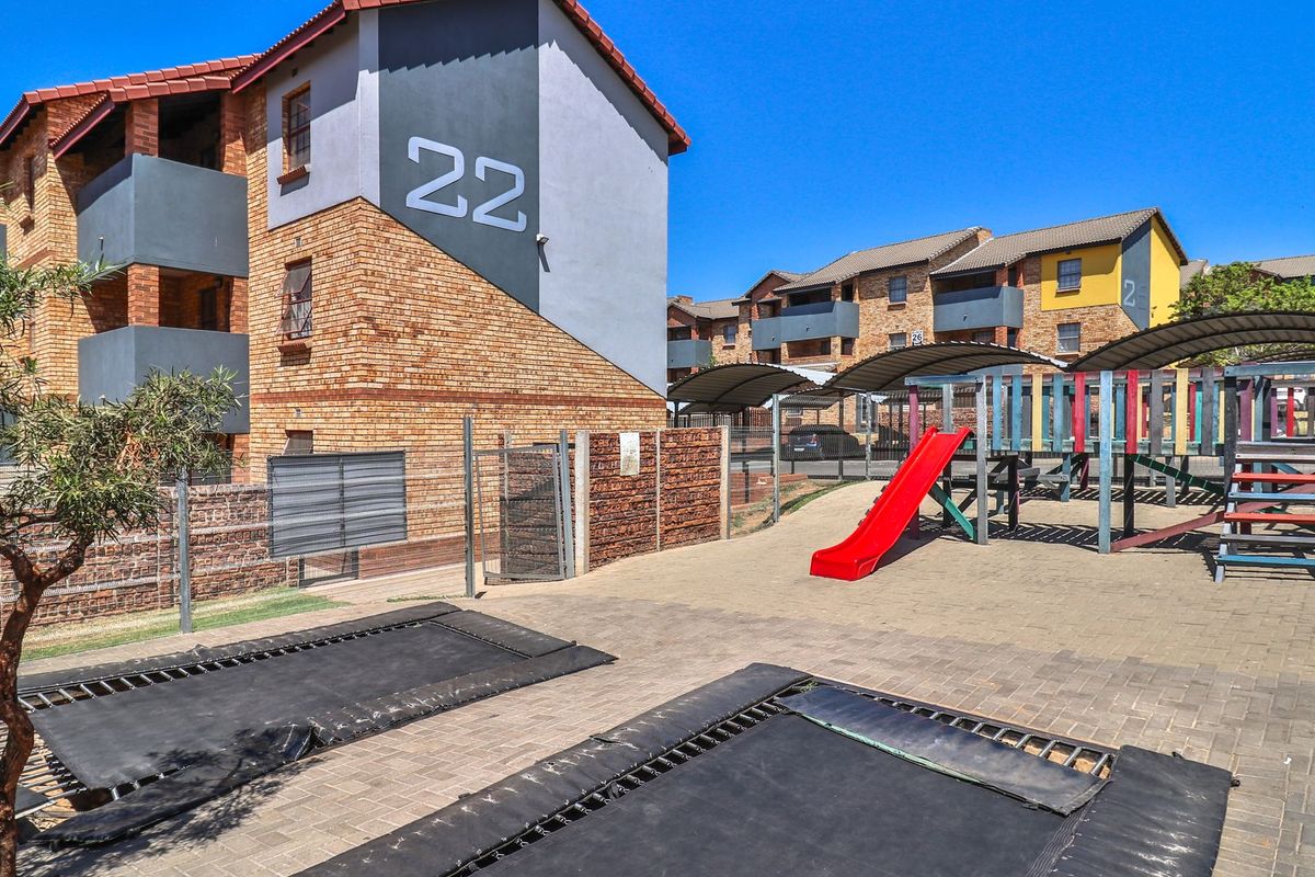 Exterior view of an apartment complex featuring a communal courtyard with a playground, in-ground trampolines, and covered parking bays.