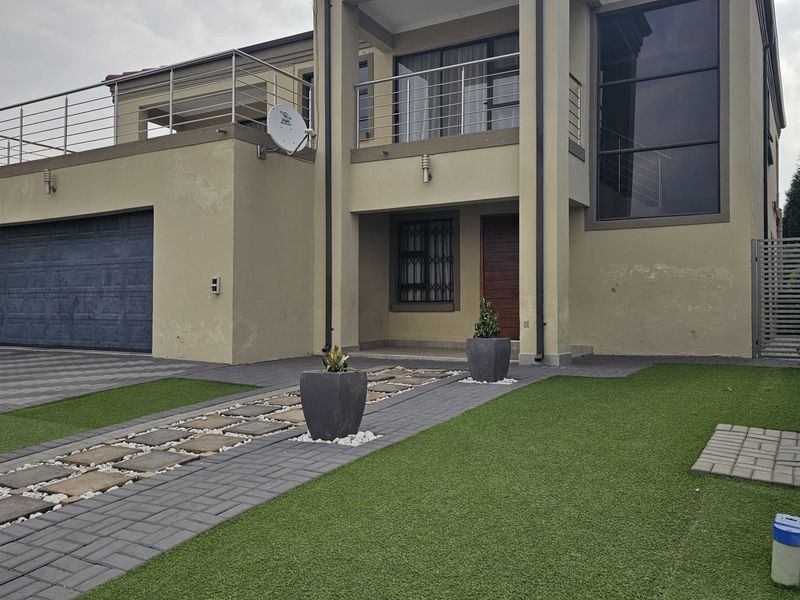 Modern double-story residential facade featuring a paved driveway, artificial lawn, integrated double garage, and an upper-level balcony with stainless steel balustrades.