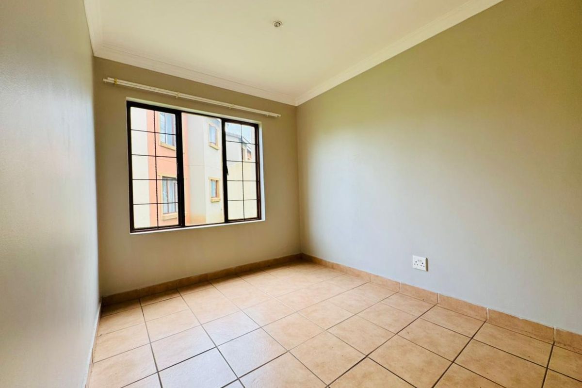 A bright, empty bedroom featuring neutral wall paint, tiled flooring, and a large window with security bars.