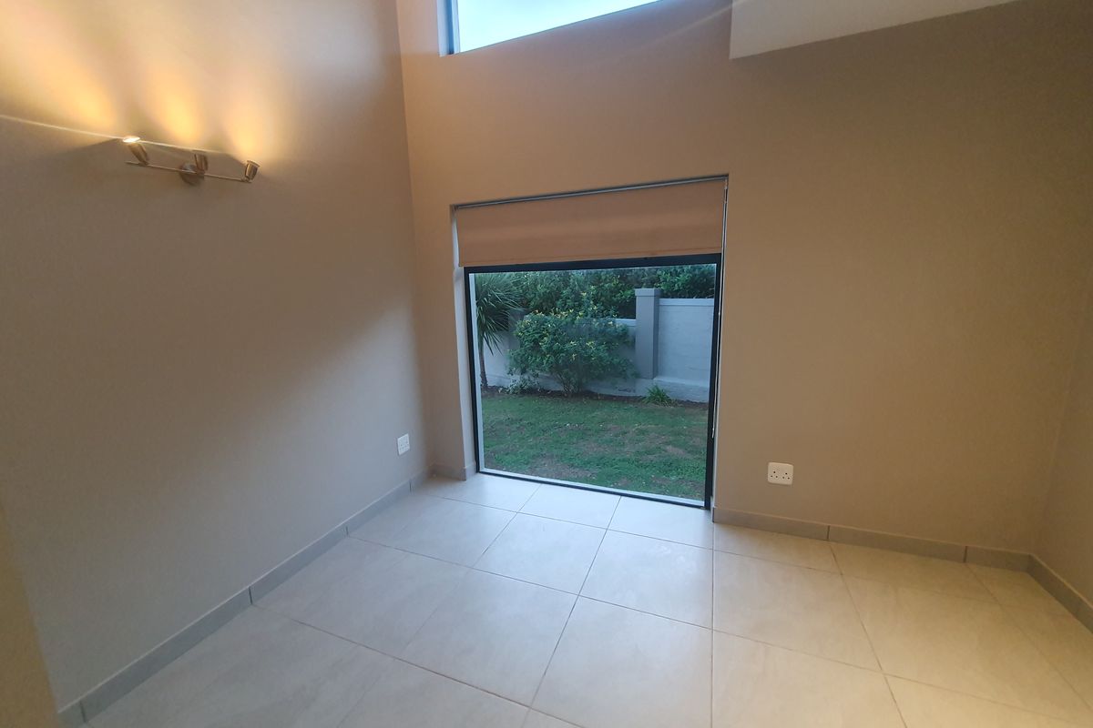 A bright, empty living space featuring high ceilings, large windows with roller blinds, tiled flooring, and modern wall-mounted lighting.