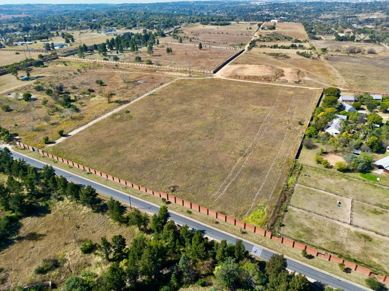 Aerial view of a large, vacant residential plot featuring perimeter walling and direct access to a paved road in a semi-rural setting.