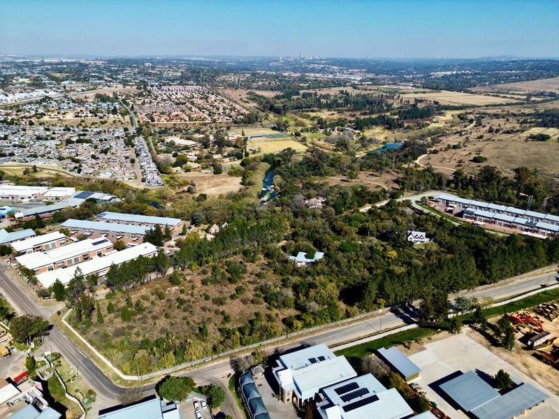 An aerial view of a vacant residential land plot situated within a developed suburban area, featuring surrounding vegetation and nearby commercial and residential infrastructure.