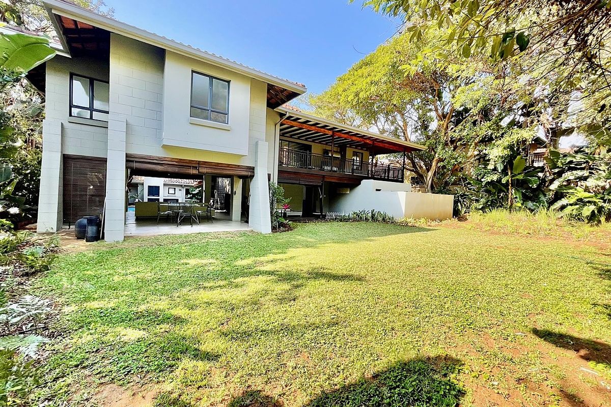 A spacious rear view of the property featuring a large landscaped garden, a covered patio area, and an elevated wooden balcony.