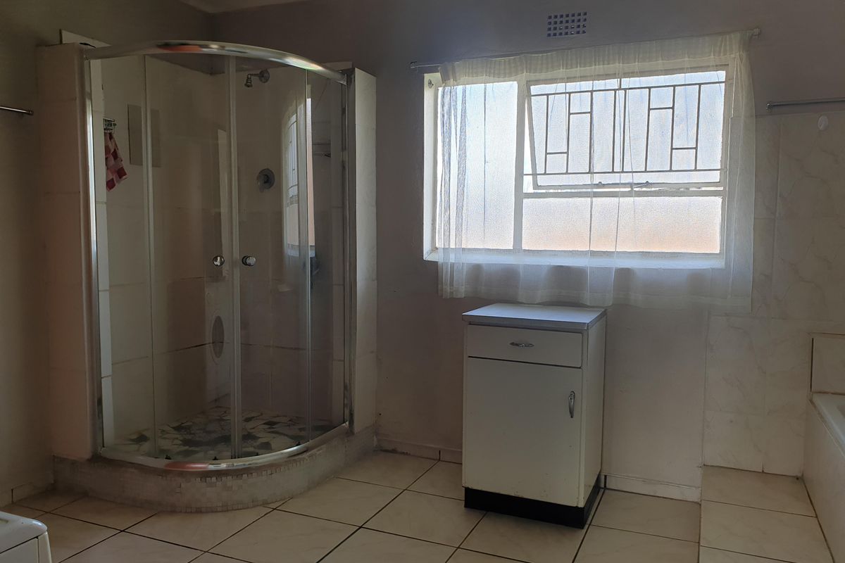 A functional bathroom featuring a curved glass shower enclosure, tiled flooring, and a window with security bars, offering potential for modernization.