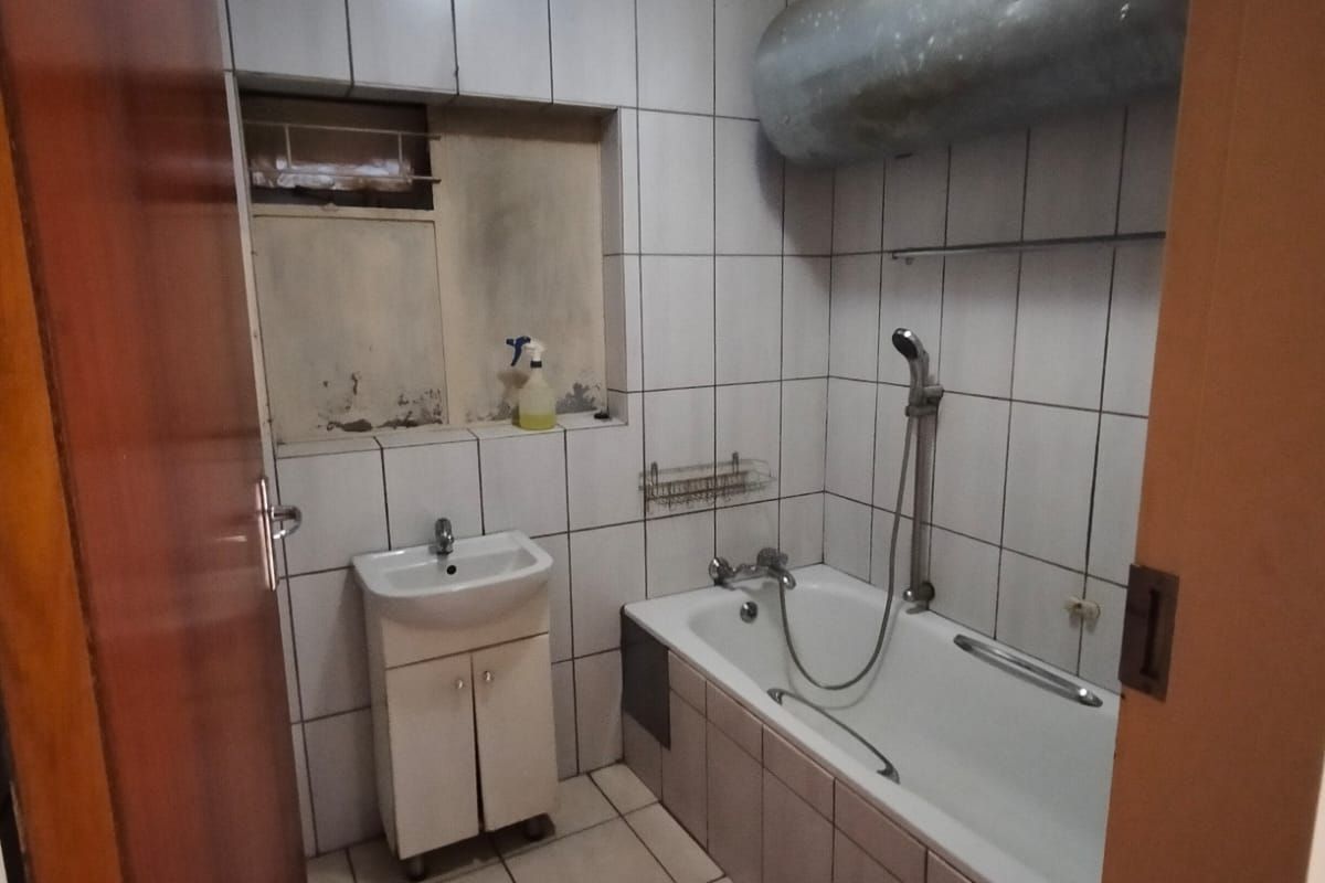A dated bathroom featuring a bathtub with shower attachment, a small vanity basin, and exposed water heating infrastructure.