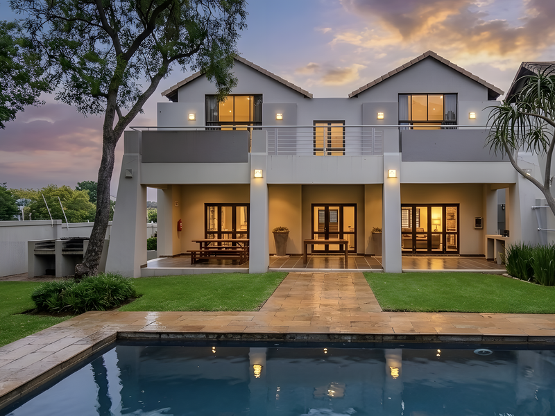 Rear view of a modern double-story apartment featuring a swimming pool, paved patio, balcony, and landscaped garden area.