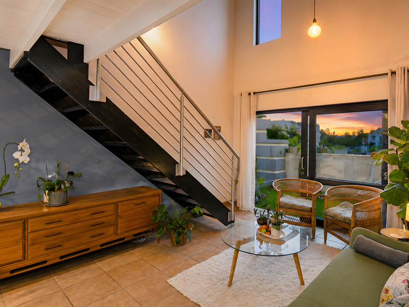 A bright living room featuring a modern staircase with stainless steel balustrades, exposed timber ceiling beams, and large sliding glass doors leading to the exterior.