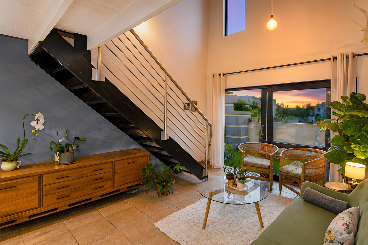 A bright living room featuring a modern staircase with stainless steel balustrades, exposed timber ceiling beams, and large sliding glass doors leading to the exterior.