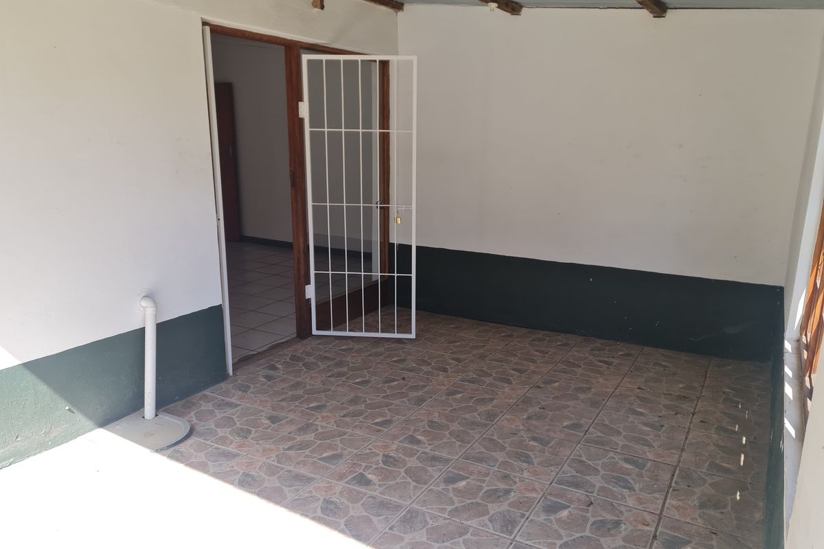 A covered outdoor patio area featuring tiled flooring, exposed timber rafters, and a security gate providing access to the interior of the flat.
