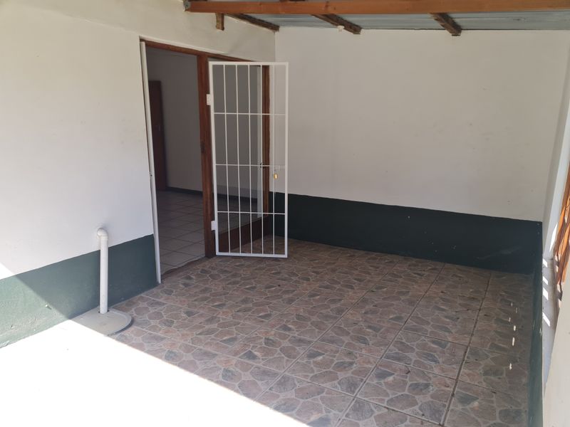 A covered outdoor patio area featuring tiled flooring, exposed timber rafters, and a security gate providing access to the interior of the flat.