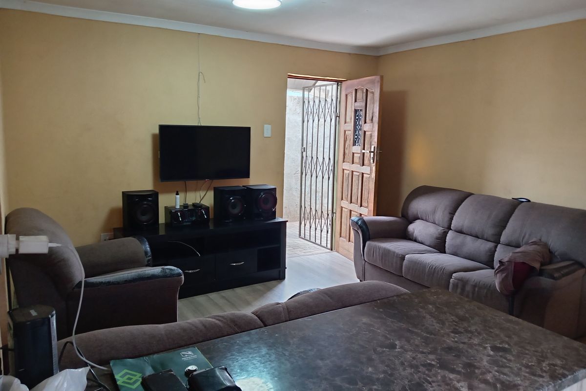 A living room area featuring a security gate, timber door, and basic ceiling lighting, with visible wear on the wall surfaces.