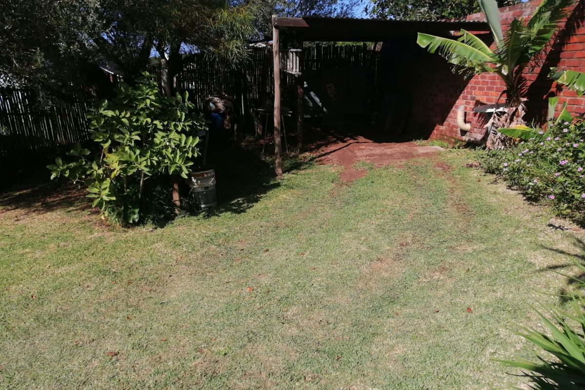A rear garden area featuring a lawn, mature trees, and a basic covered outdoor structure against a brick boundary wall.