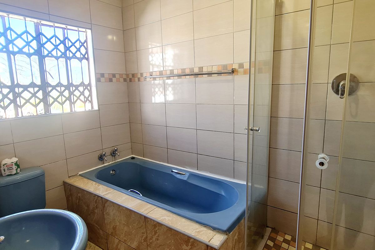 A functional bathroom featuring a blue bathtub, matching basin, and a separate glass-enclosed shower with neutral wall tiling and security bars on the window.