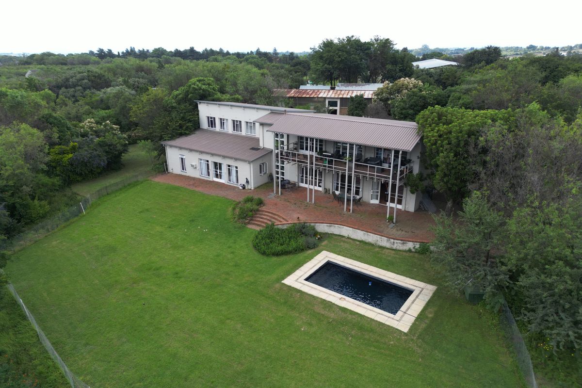 Aerial view of a two-story residence featuring a swimming pool, expansive lawn, covered patio, and upper-level balcony set within a lush, green landscape.
