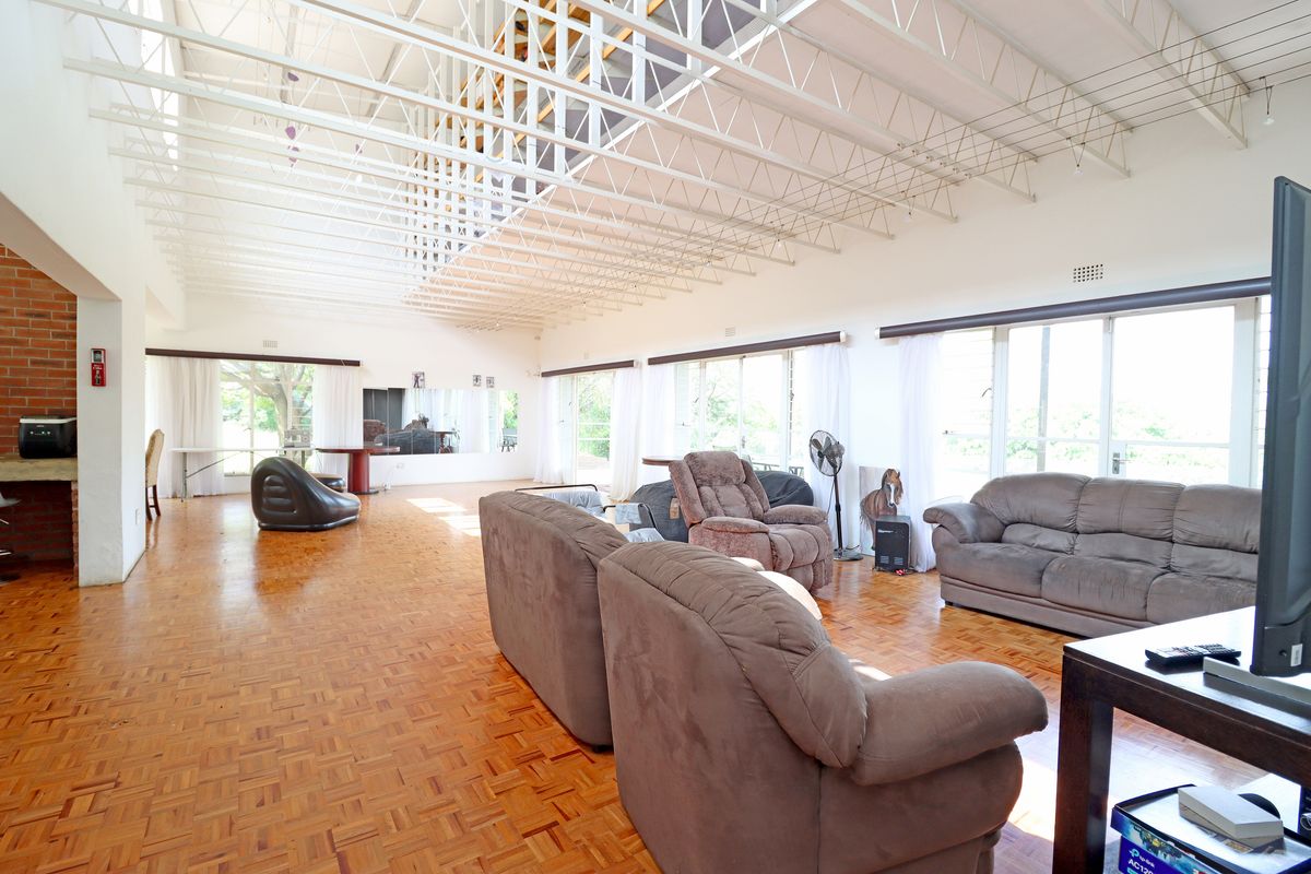 Spacious living area featuring high ceilings with exposed steel trusses, parquet flooring, and a brick feature wall with large windows providing natural light.
