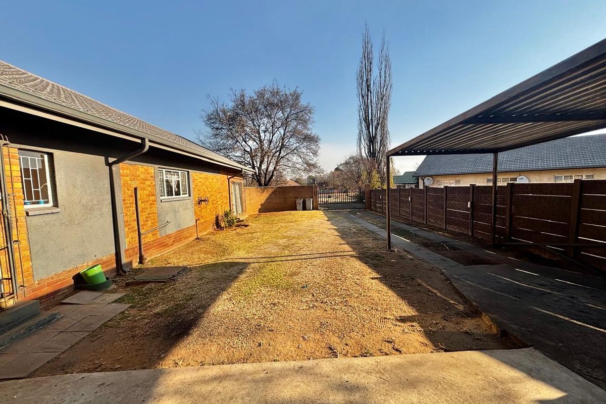A rear exterior view of the property featuring a lawn area, a metal carport structure, and perimeter boundary walls.