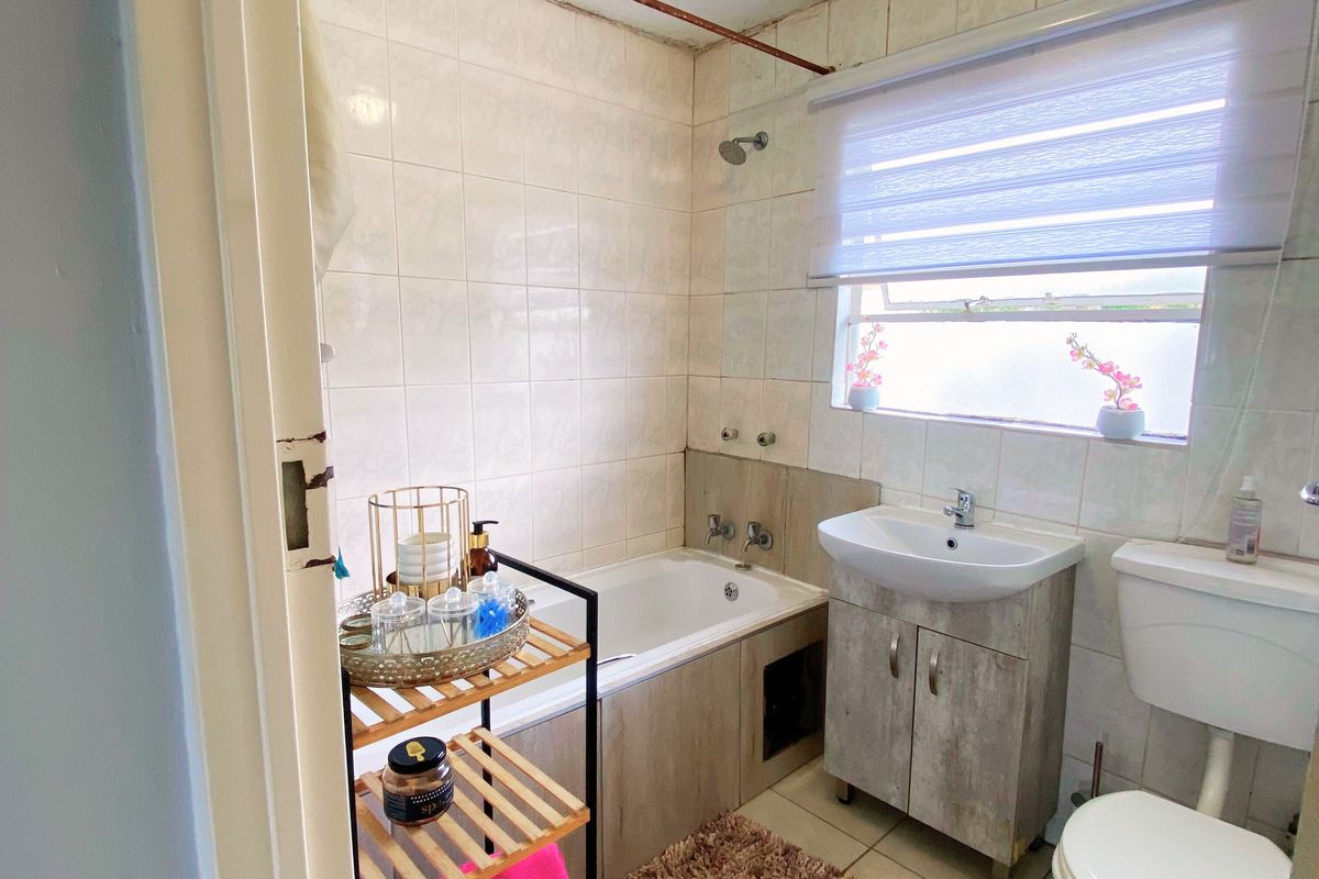 A functional bathroom featuring a built-in bathtub with shower mixer, vanity basin, and toilet, finished with neutral wall and floor tiling.