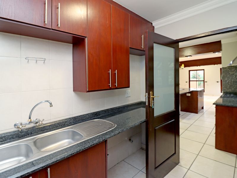 A functional scullery area featuring dark wood cabinetry, granite countertops, a double sink, and tiled finishes, with direct access to the main kitchen area.