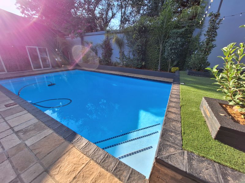 A well-maintained swimming pool area featuring stone paving, artificial turf, and raised garden beds against a perimeter wall.