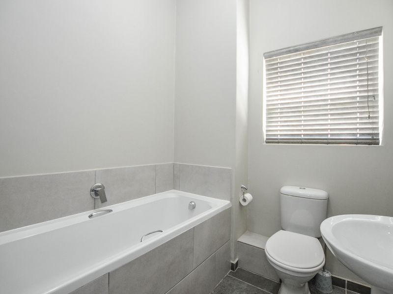 A clean and functional bathroom featuring a built-in bathtub, pedestal washbasin, toilet, and neutral wall tiling with natural light from a window.