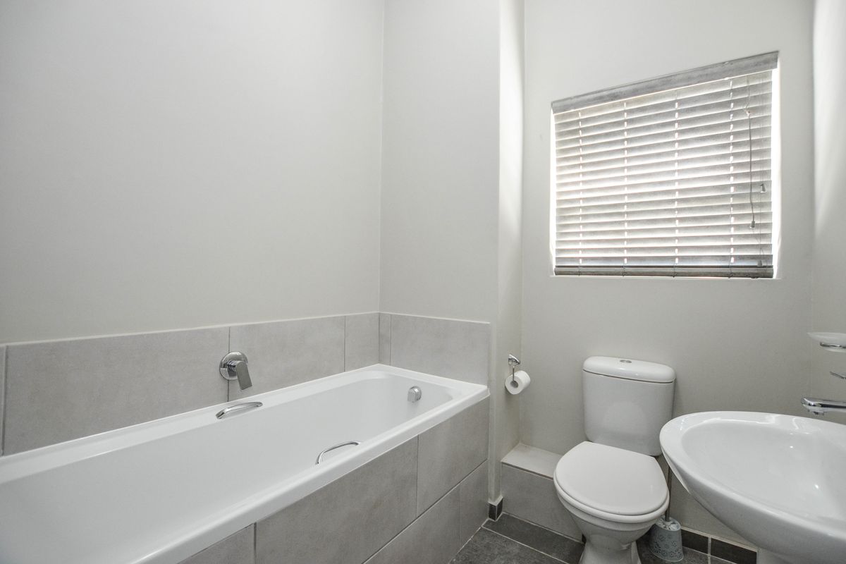 A clean and functional bathroom featuring a built-in bathtub, pedestal washbasin, toilet, and neutral wall tiling with natural light from a window.