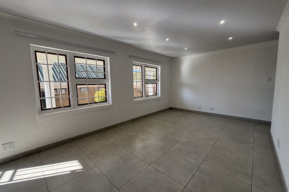 A bright, empty living room featuring neutral tiled flooring, recessed downlighting, and large windows fitted with security burglar bars.
