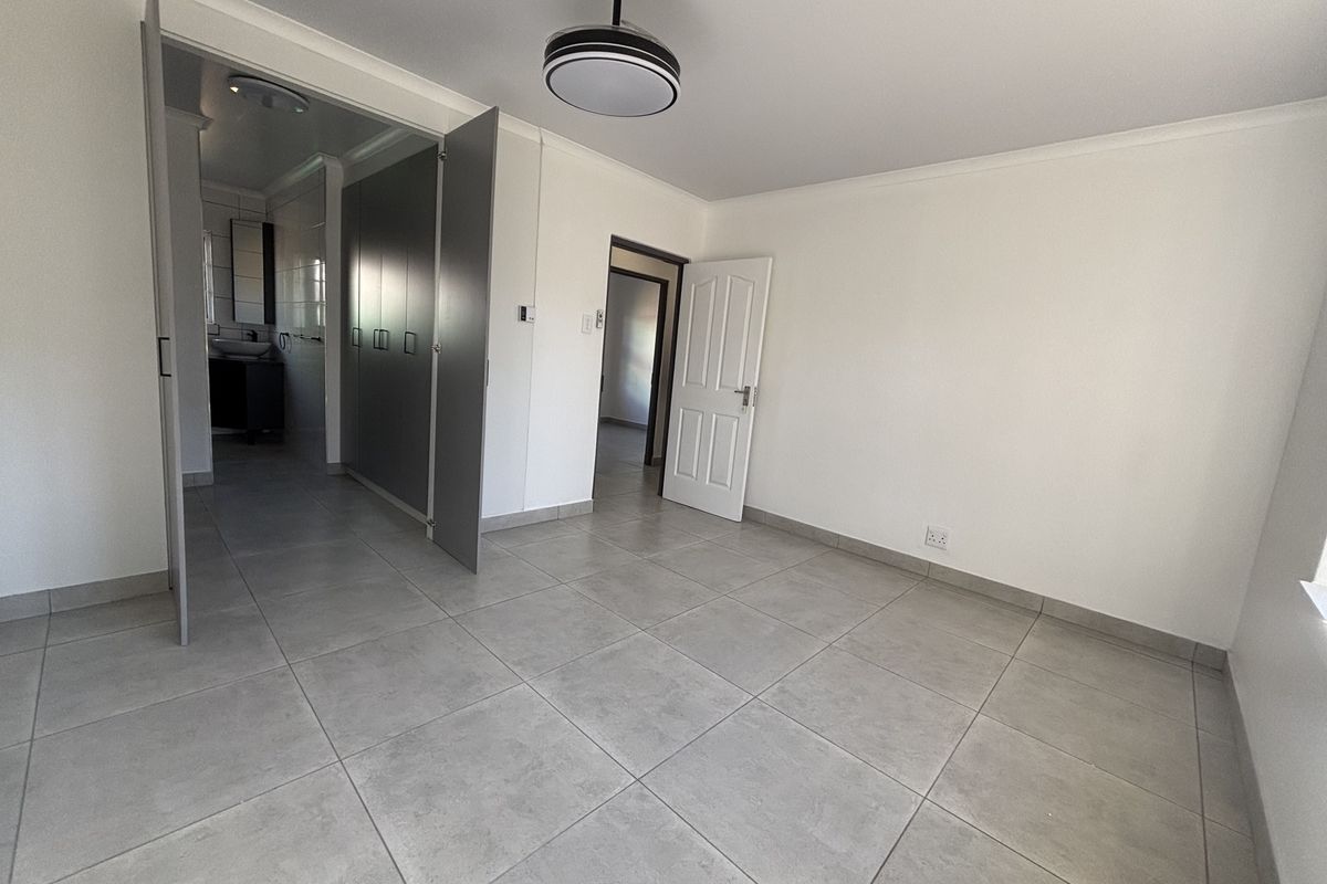 A spacious, empty bedroom featuring neutral tiled flooring, built-in cupboards, and a modern ceiling fan, with direct access to an en-suite bathroom.