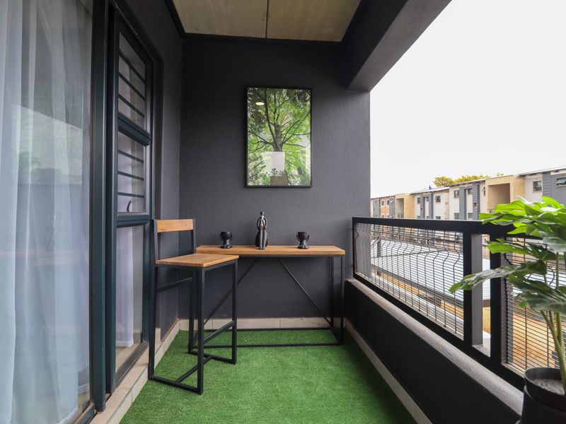 A modern covered balcony featuring dark grey walls, artificial grass flooring, and a metal balustrade, providing a compact outdoor space.