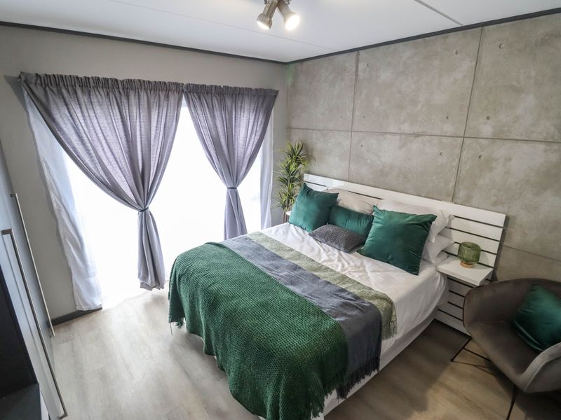 A modern bedroom featuring a concrete-look feature wall, light laminate flooring, and a built-in wardrobe with ample natural light from the window.