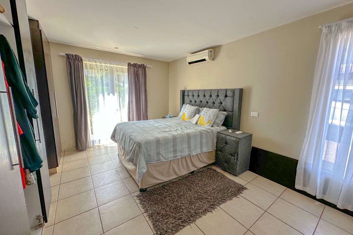 A spacious bedroom featuring tiled flooring, a wall-mounted air conditioning unit, and large windows with curtains providing natural light.