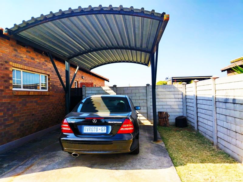 A covered carport area featuring a corrugated metal roof, concrete driveway, and secure precast concrete boundary walls.
