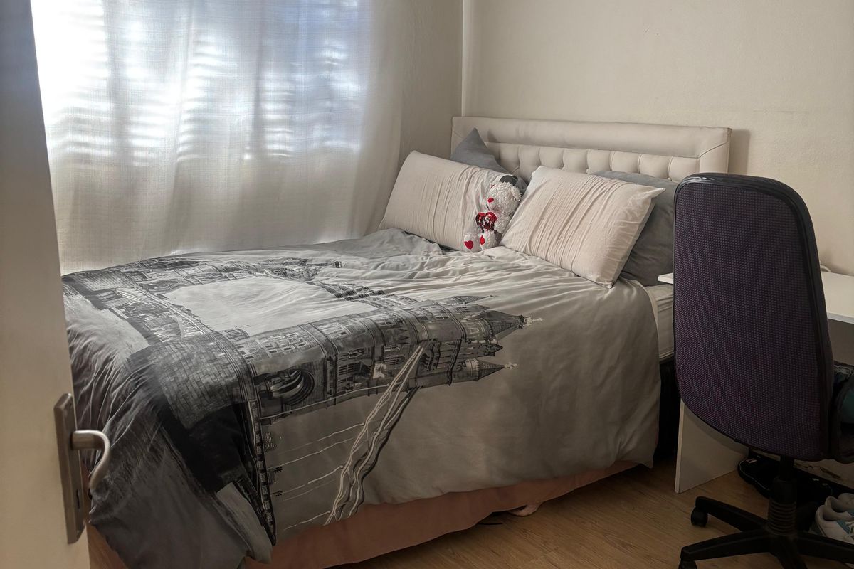 A compact bedroom featuring light wood-look laminate flooring and a window with sheer curtains.