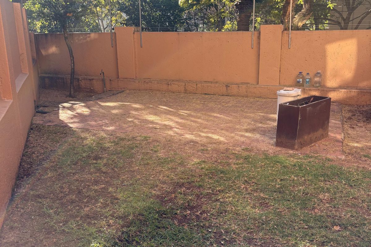 A private rear garden area featuring a paved section, lawn, and high perimeter walls with security fencing.