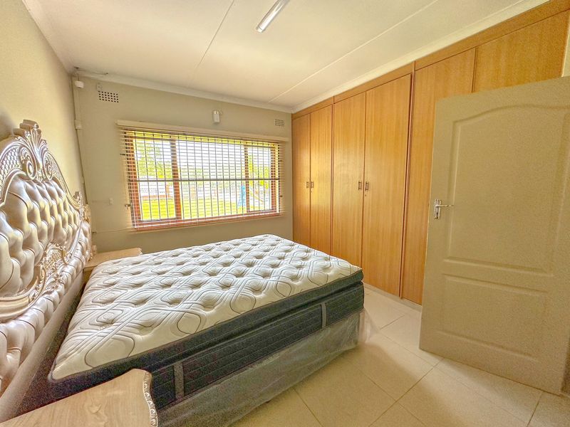 A bedroom featuring built-in wooden cupboards, tiled flooring, and a window fitted with security bars and venetian blinds.