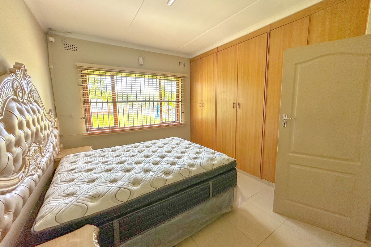A bedroom featuring built-in wooden cupboards, tiled flooring, and a window fitted with security bars and venetian blinds.