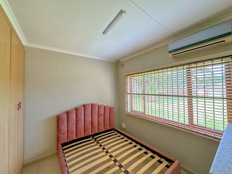 A well-maintained bedroom featuring built-in wardrobes, tiled flooring, and a wall-mounted air conditioning unit, with natural light provided by a large window.