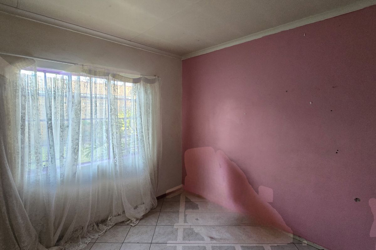 An empty bedroom featuring tiled flooring and a window with security bars, currently requiring wall repairs and repainting to improve its condition.