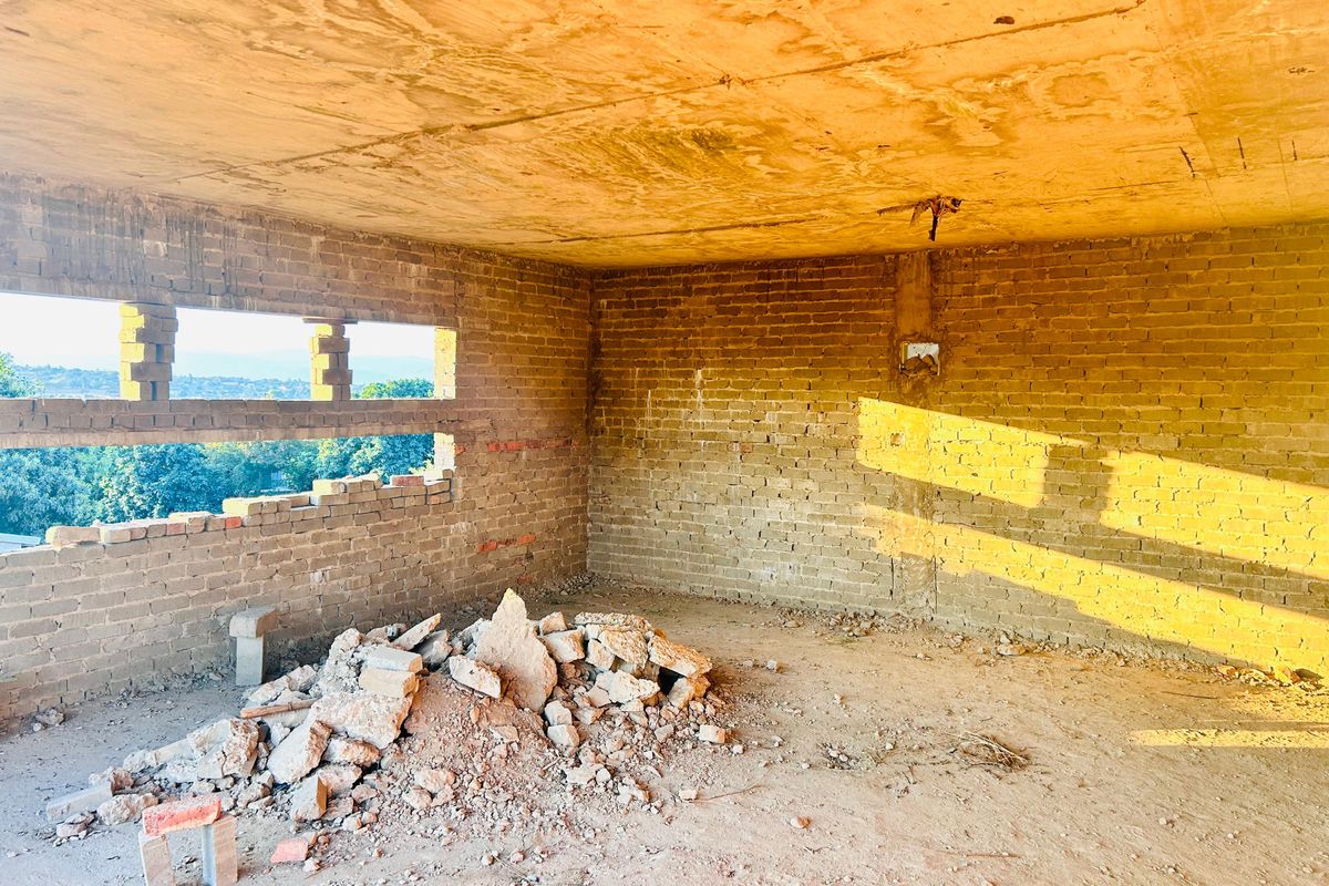 An unfinished bedroom space featuring exposed brick walls, a concrete ceiling, and horizontal window openings with views of the surrounding landscape.