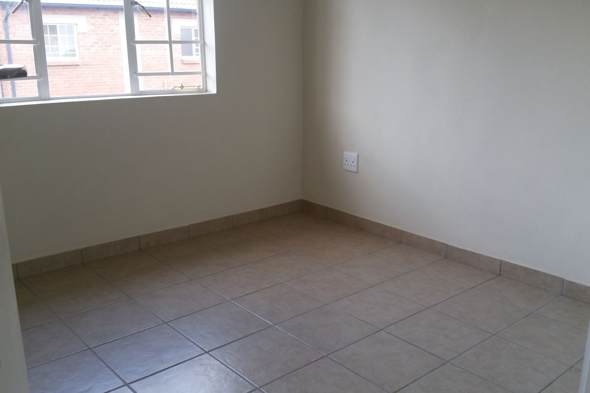 A clean, empty bedroom featuring neutral ceramic floor tiles, a window with security bars, and a standard electrical outlet.
