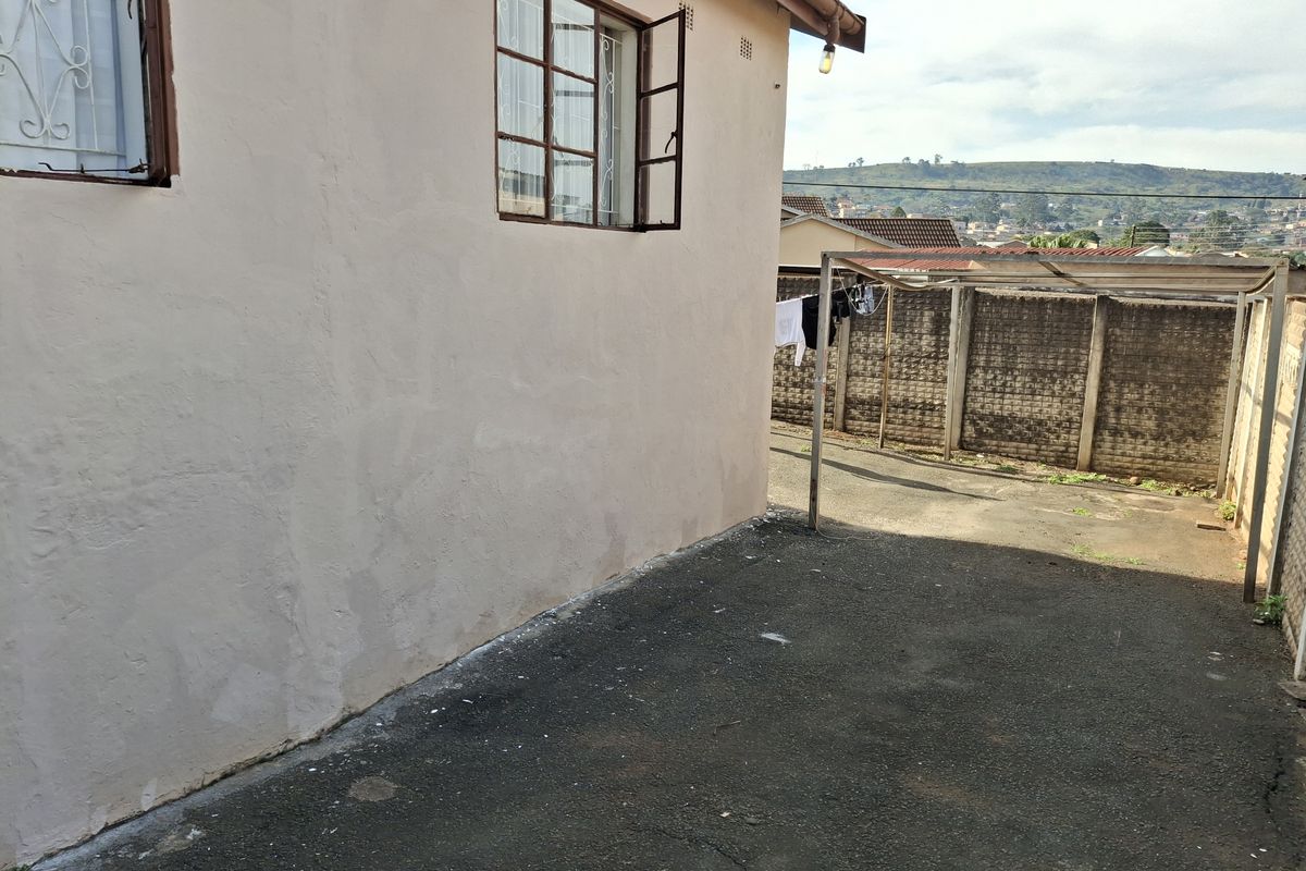 View of the rear exterior area featuring a paved driveway, perimeter boundary wall, and metal structure, showing signs of wear and weathering.