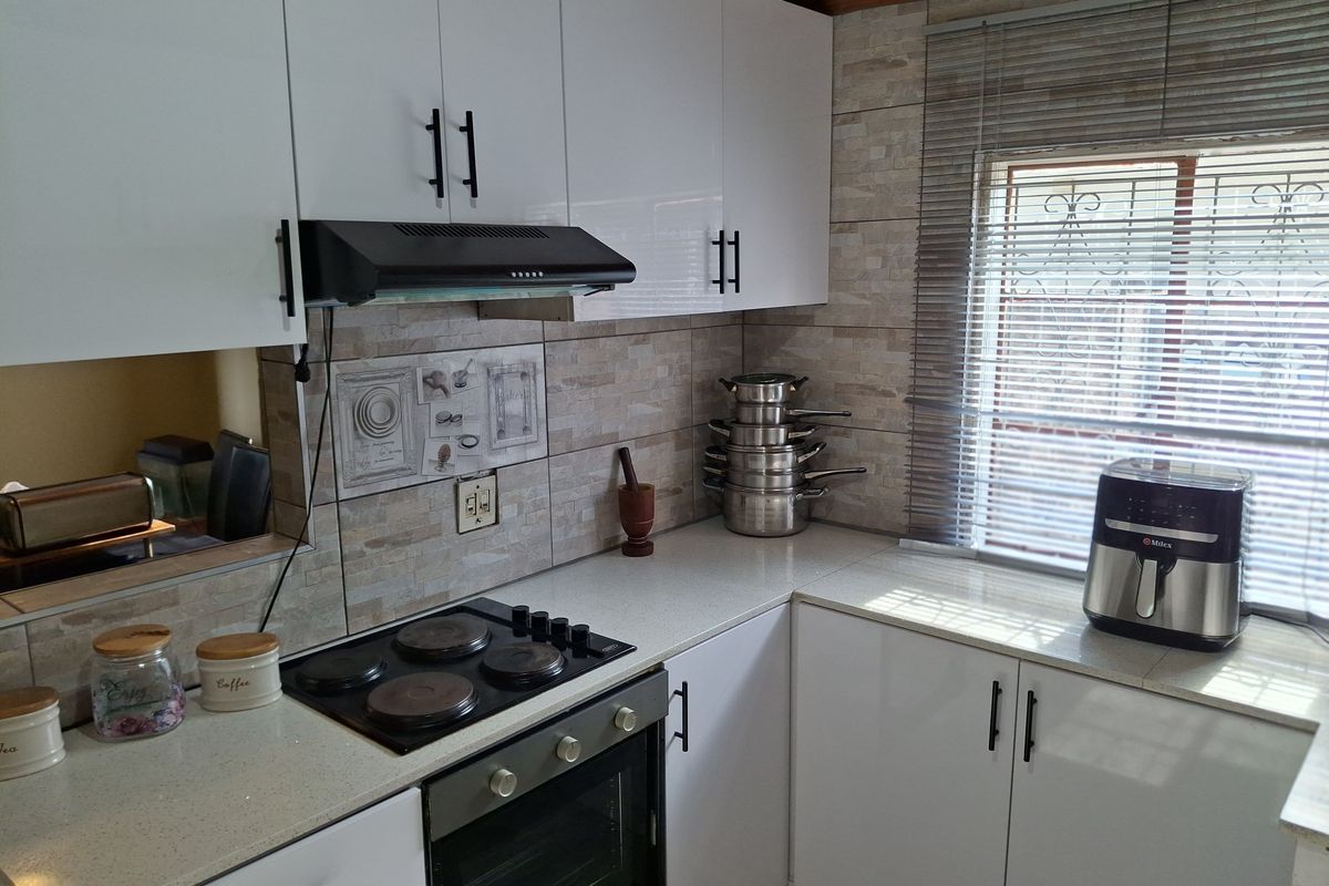 A functional kitchen featuring white cabinetry, stone-look countertops, a built-in electric hob, oven, and extractor fan, complemented by tiled splashbacks and a window with blinds.