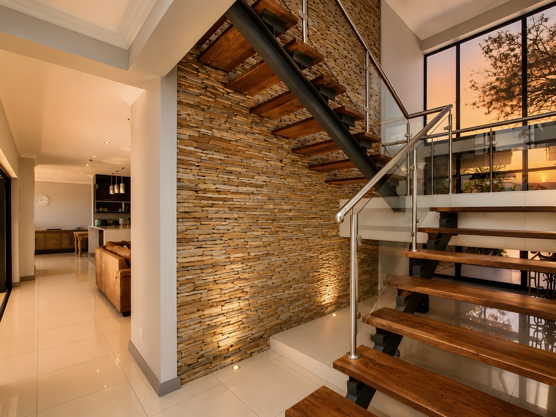 Elegant entrance hall featuring a striking stone feature wall, modern timber staircase with glass balustrade, and large windows overlooking the exterior.