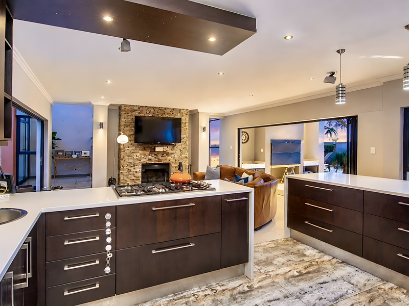 Modern open-plan kitchen and living area featuring dark wood cabinetry, stone countertops, a gas hob, and a stone-clad fireplace with seamless outdoor flow.