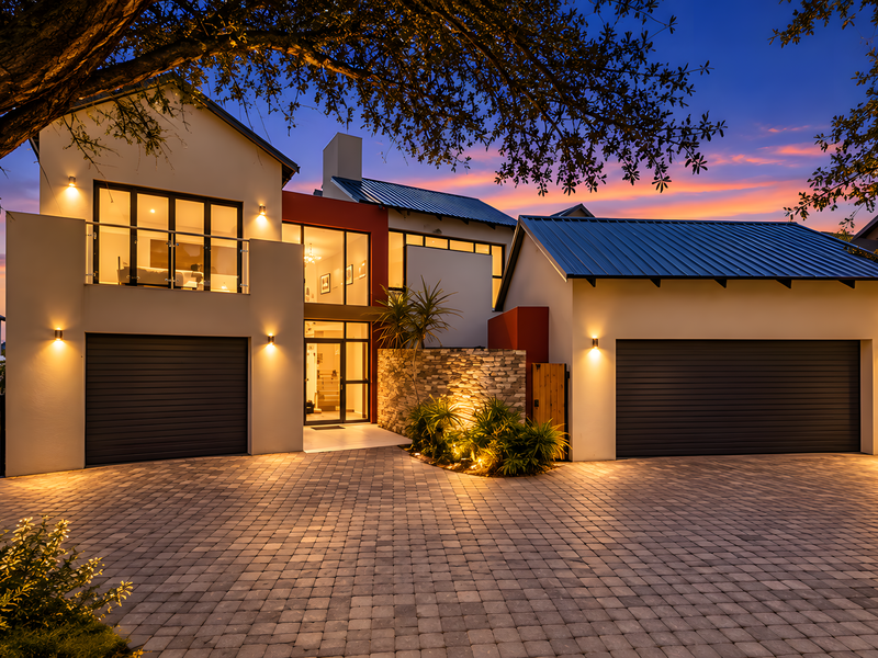 Modern double-story residence featuring a paved driveway, double garage, stone-clad accents, and a balcony, illuminated by ambient exterior lighting at dusk.