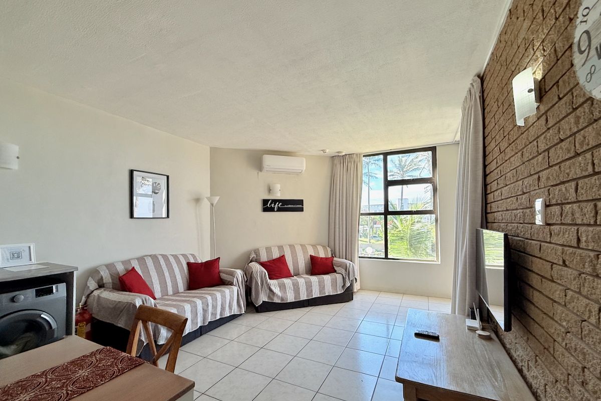 A bright living room featuring tiled flooring, an exposed brick feature wall, and a split-system air conditioning unit with natural light from a large window.