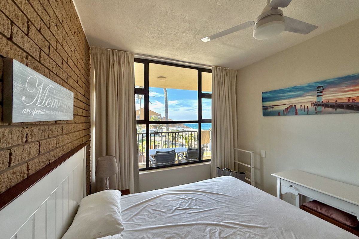 A bright bedroom featuring an exposed brick feature wall, ceiling fan, and large window with views of the balcony and ocean.