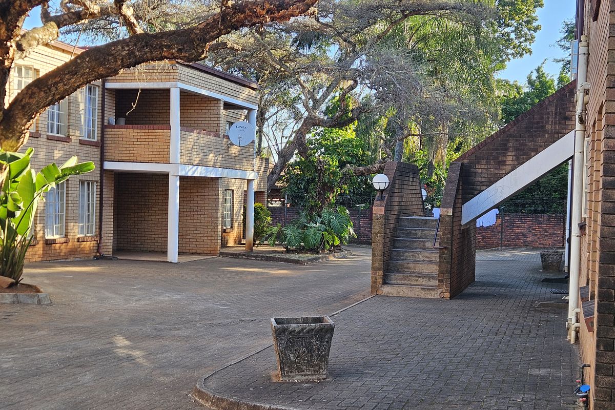 Exterior view of an apartment complex courtyard featuring face-brick architecture, paved walkways, external staircases, and mature trees in a communal garden setting.
