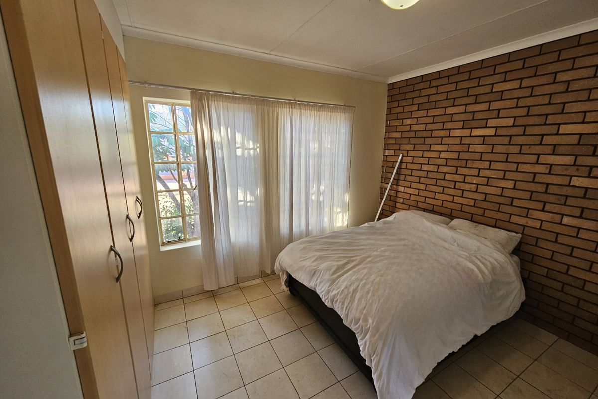 A bedroom featuring a prominent face-brick feature wall, built-in wooden cupboards, tiled flooring, and a window with natural light.