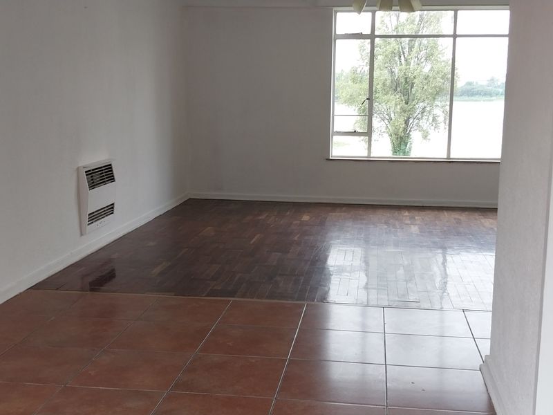 An empty living room featuring a combination of ceramic tiled and parquet flooring, a wall-mounted heater, and a large window with natural light.